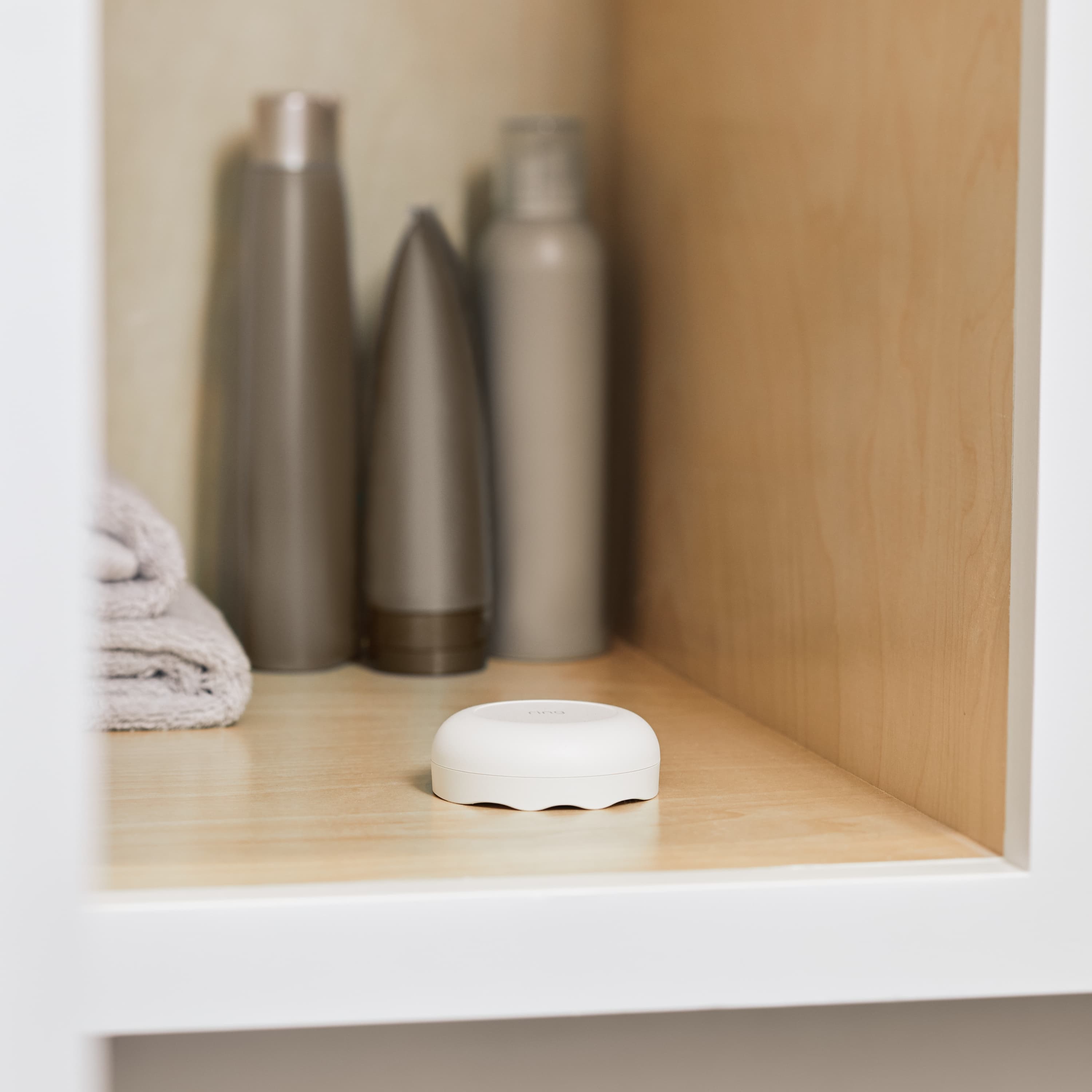 Flood & Freeze Alarm Bundle - White Ring Alarm Flood and Freeze Sensor placed on a wooden shelf next to folded towels and toiletry bottles. 