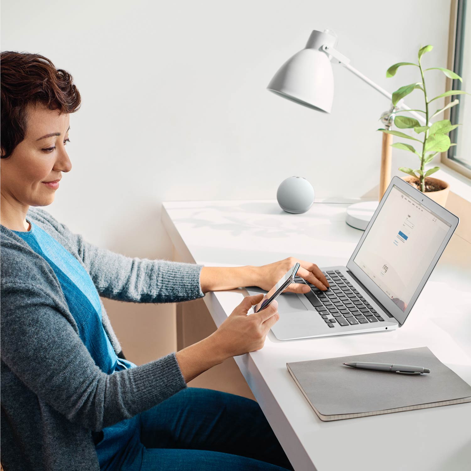 Alarm Pro Security Kit, 14-Piece (with built-in eero Wi-Fi 6 router) - Woman sitting at desk using a laptop and smartphone, with a smart speaker, desk lamp, and potted plant in background.