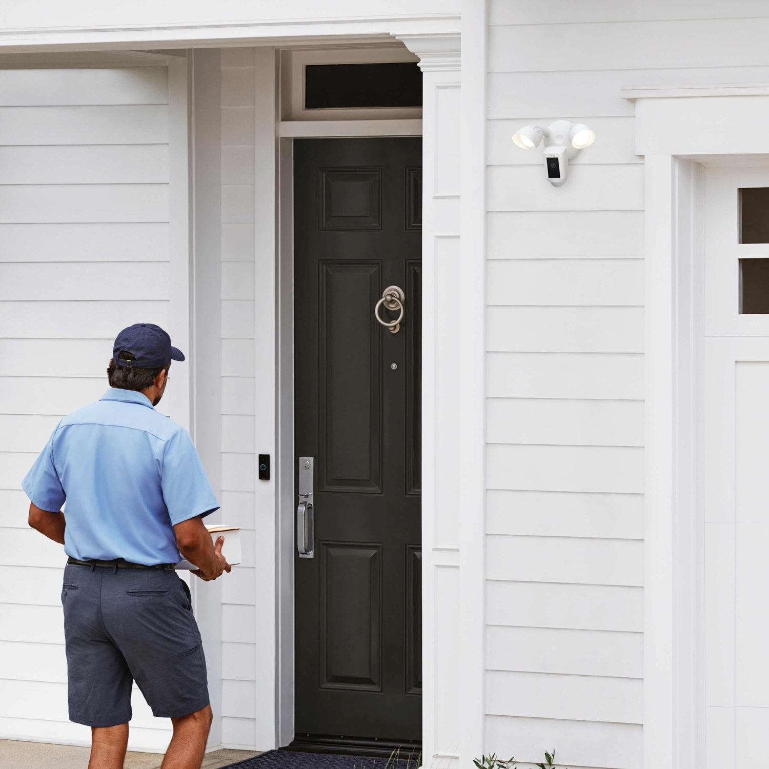 Floodlight Cam Wired Plus (Certified Refurbished) - Delivery driver approaches front door with package on ground, while Ring Floodlight Cam Wired Plus monitors from above.