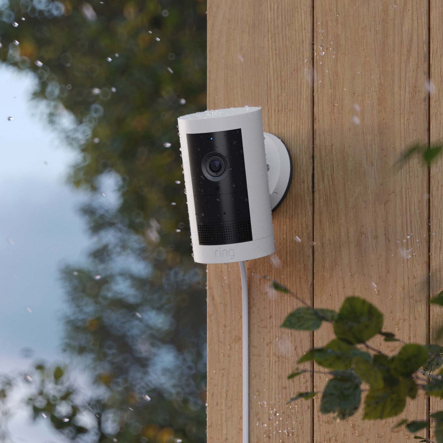 Outdoor Cam Plus Plug-In + Indoor Cam (2nd Gen) - Plug-In:White Ring Outdoor Cam Plus mounted on wood wall, covered in raindrops, with rain falling.