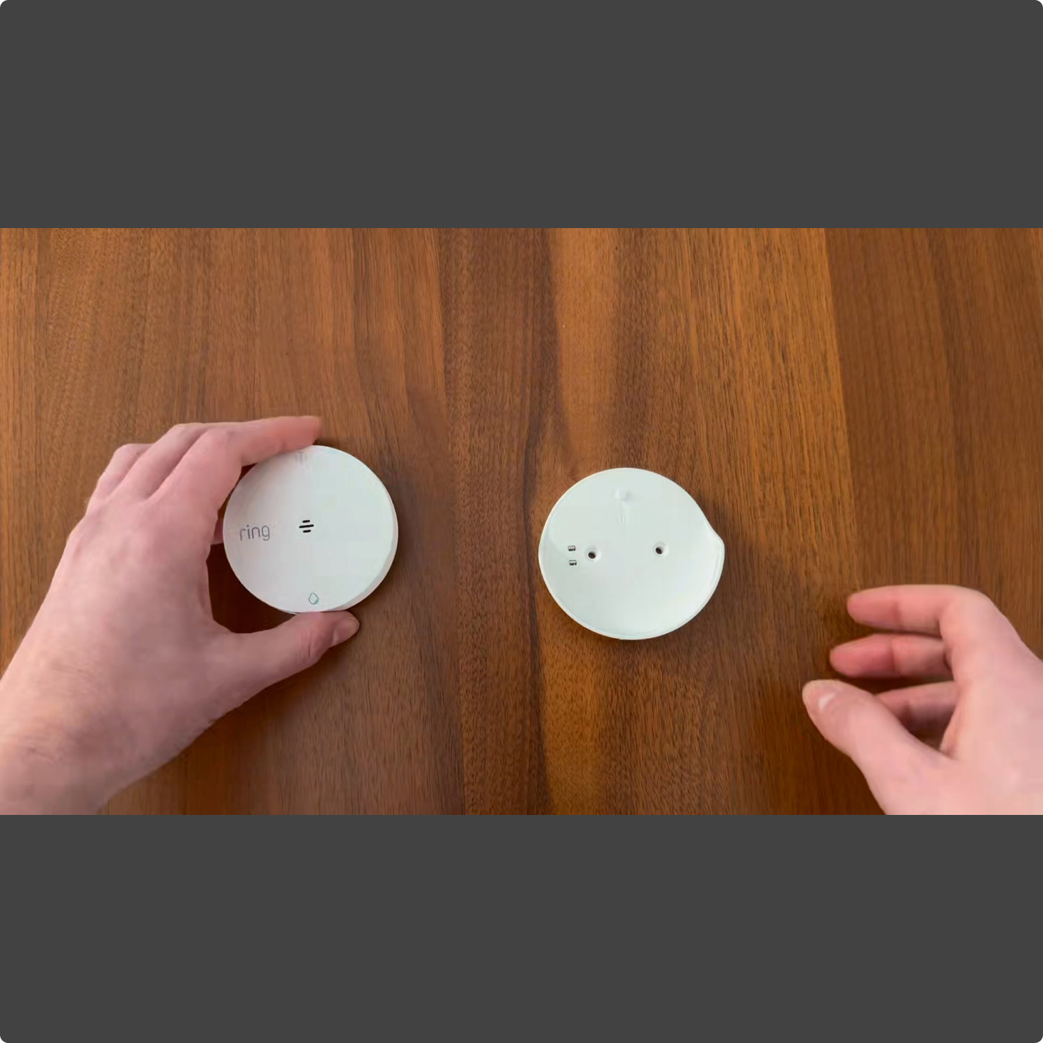 Sump Pump Monitor (Ring Sensors) - Hands hold two small white round devices on a wooden table, one labeled “ring,” as if preparing to install or compare them.