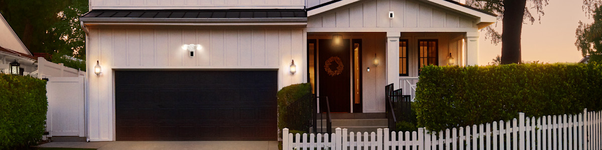 Modern white farmhouse with black garage door and white picket fence at dusk displaying a Solar Security Sign